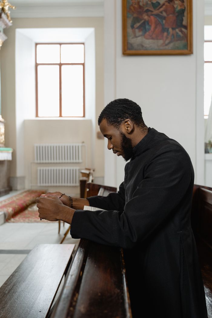 services-01 Side view of a bearded African American priest praying in a church pew.